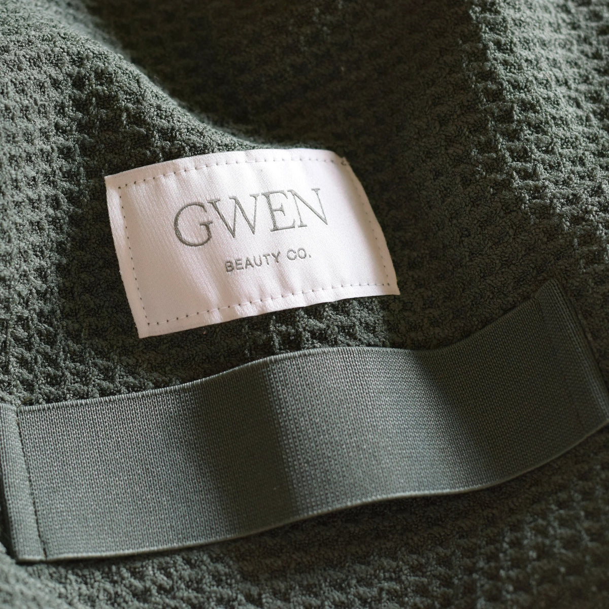 The GWEN Hair Towel in Moss, the limited-edition colorway, shown as a flat lay with the woven GWEN Beauty Co. label visible.